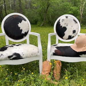 May include: Two white armchairs with cowhide seats and backs, accented with gold studs. A tan hat rests on one chair, and a pair of brown cowboy boots sits in front of the chairs. The chairs are in a field of yellow flowers.