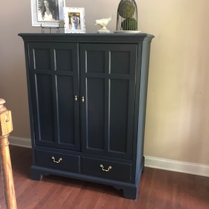 May include: A navy blue wooden cabinet with two doors and two drawers. The cabinet has gold-colored hardware and decorative molding. The top of the cabinet displays framed photos and decorative objects.