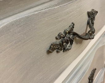 May include: Close-up of a painted dresser with ornate bronze handles. The dresser has a light gray finish with white and light blue accents. The handles are detailed with floral designs.
