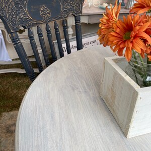 May include: A distressed black wooden chair with a carved back and a white painted round table with a wood grain pattern. A white wooden box and a glass jar with orange flowers are on the table.