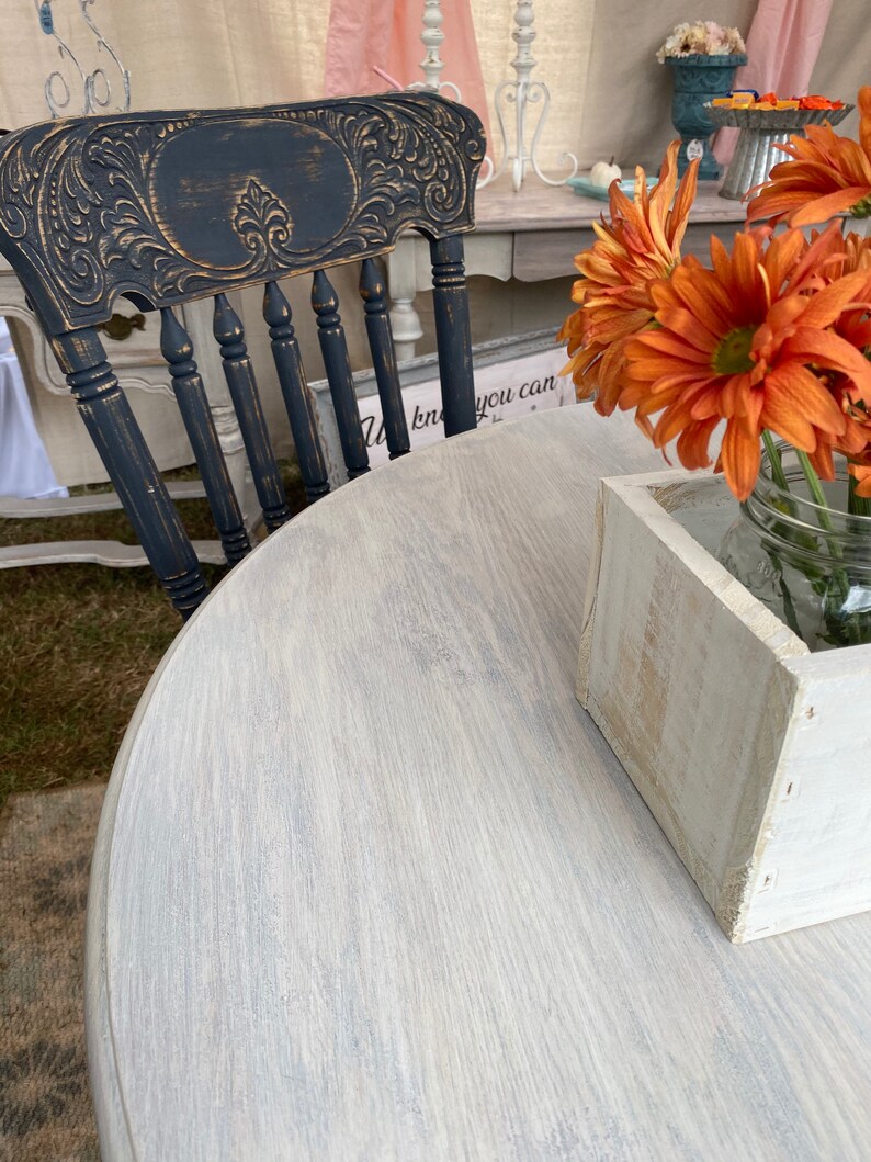 May include: A distressed black wooden chair with a carved back and a white painted round table with a wood grain pattern. A white wooden box and a glass jar with orange flowers are on the table.