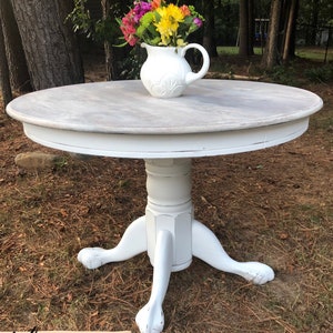 May include: White round table with a distressed white finish and a pedestal base with three legs. The table is decorated with a bouquet of colorful flowers in a white pitcher.
