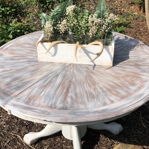 Puede incluir: Una mesa redonda blanca con una tapa de madera desgastada y una base de pedestal blanca. La mesa est&aacute; decorada con una caja de madera blanca llena de vegetaci&oacute;n y cuernos.