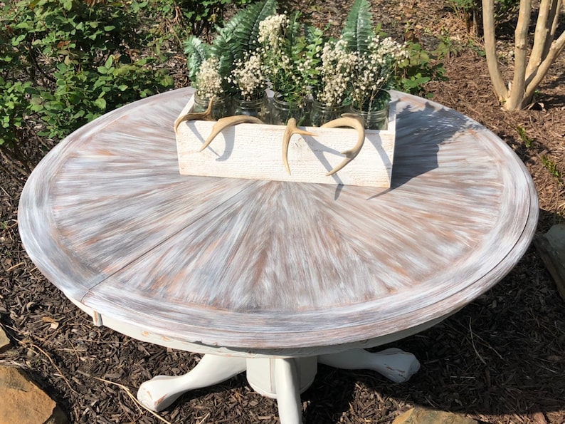 Puede incluir: Una mesa redonda blanca con una tapa de madera desgastada y una base de pedestal blanca. La mesa est&aacute; decorada con una caja de madera blanca llena de vegetaci&oacute;n y cuernos.