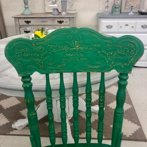 Pu&ograve; includere: Una sedia in legno verde con schienale intagliato e gambe a fuso. La sedia &egrave; dipinta di verde scuro con accenti dorati. La sedia &egrave; appoggiata su un tappeto bianco e grigio.