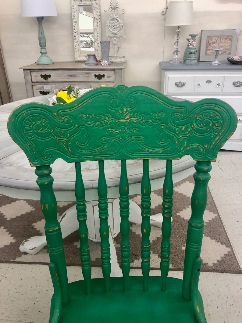 Pu&ograve; includere: Una sedia in legno verde con schienale intagliato e gambe a fuso. La sedia &egrave; dipinta di verde scuro con accenti dorati. La sedia &egrave; appoggiata su un tappeto bianco e grigio.