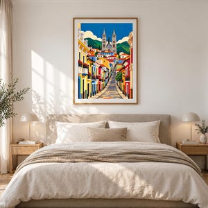 May include: A framed vertical art print featuring a vibrant street scene with colorful buildings leading toward a large cathedral under a bright blue sky with white clouds, set against a backdrop of green mountains and rolling hills.