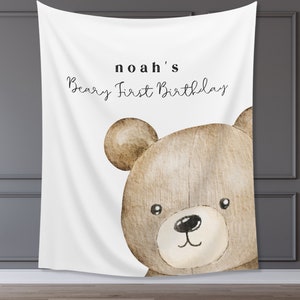 Może przedstawiać: Biały baner z tkaniny z ilustracją brązowego misia. Na banerze widnieje napis "noah's Beary First Birthday" czarnym skryptem. Miś ma czarny nos i małe czarne oczy. Idealny na pierwsze urodziny.