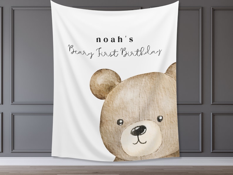 Może przedstawiać: Biały baner z tkaniny z ilustracją brązowego misia. Na banerze widnieje napis "noah's Beary First Birthday" czarnym skryptem. Miś ma czarny nos i małe czarne oczy. Idealny na pierwsze urodziny.