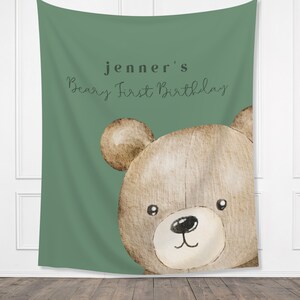 Może przedstawiać: Zielony gobelin z akwarelową ilustracją twarzy misia. Tekst "jenner's Beary First Birthday" jest napisany czcionką skryptową. Miś ma brązowe futro i czarny nos.