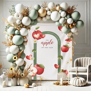 Puede incluir: Decoraci&oacute;n de baby shower con un arco de globos en blanco, verde salvia y dorado. El tel&oacute;n de fondo incluye un letrero con el texto "hannah's baby shower" y "the apple of our eye" con ilustraciones de manzanas y corazones. Las calabazas y flores decorativas completan la escena.