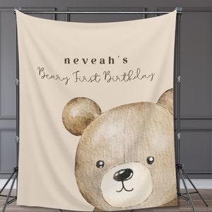 Może przedstawiać: Beżowe tło z akwarelową ilustracją głowy misia. Tekst "neveah's Beary First Birthday" jest napisany wielkimi i małymi literami. Tło jest podparte czarnym stojakiem.