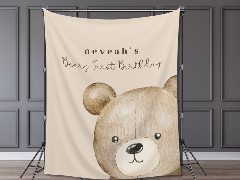 Może przedstawiać: Beżowe tło z akwarelową ilustracją głowy misia. Tekst "neveah's Beary First Birthday" jest napisany wielkimi i małymi literami. Tło jest podparte czarnym stojakiem.