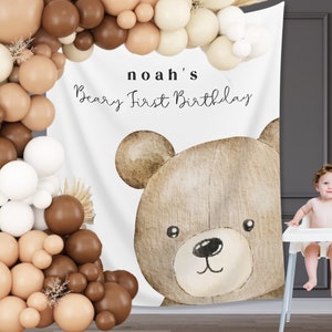 Może przedstawiać: Tło na przyjęcie urodzinowe z dużą akwarelową ilustracją misia i napisem "noah's Beary First Birthday". Łuk balonowy w odcieniach brązu i kremu obramowuje tło. Dziecko siedzi w krzesełku do karmienia.
