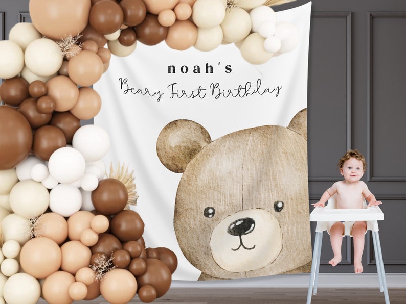 Może przedstawiać: Tło na przyjęcie urodzinowe z dużą akwarelową ilustracją misia i napisem "noah's Beary First Birthday". Łuk balonowy w odcieniach brązu i kremu obramowuje tło. Dziecko siedzi w krzesełku do karmienia.