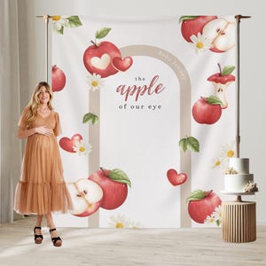 Puede incluir: Una persona embarazada con un vestido marr&oacute;n frente a un tel&oacute;n de fondo con manzanas rojas, margaritas y el texto "the apple of our eye". Un pastel blanco de dos pisos est&aacute; sobre un soporte de madera.