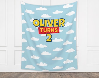 May include: A blue backdrop with white clouds and the text "OLIVER TURNS 2" in yellow and red letters. The backdrop is perfect for a birthday party.