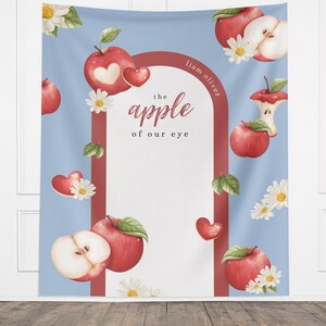 Puede incluir: Un tapiz azul con un dise&ntilde;o de acuarela con manzanas rojas, margaritas y corazones. El texto "the apple of our eye" se muestra dentro de un arco rojo. El nombre "Liam Oliver" est&aacute; escrito en el lado derecho del arco.