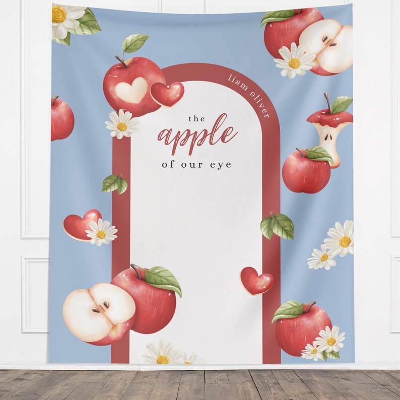 Puede incluir: Un tapiz azul con un dise&ntilde;o de acuarela con manzanas rojas, margaritas y corazones. El texto "the apple of our eye" se muestra dentro de un arco rojo. El nombre "Liam Oliver" est&aacute; escrito en el lado derecho del arco.