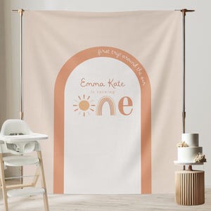 Puede incluir: Un telón de fondo de color melocotón con un gran diseño de arco en naranja y blanco. El arco presenta un sol, un arcoíris y la palabra "uno" en una fuente caprichosa. El texto "primer viaje alrededor del sol" y "Emma Kate está cumpliendo" también está incluido. Este telón de fondo es perfecto para una fiesta de primer cumpleaños.
