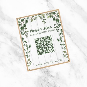 Puede incluir: Un cartel de madera blanco con un borde de hojas verdes y un código QR en el centro. El texto en el cartel dice "Kenzie & Jake's Honeymoon Fund" y "¡Muchas gracias!"