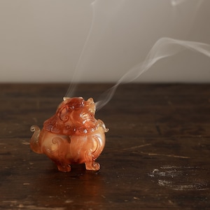May include: A small, amber-colored incense burner shaped like a mythical creature with intricate details. Smoke curls up from the burner, which is sitting on a dark wooden surface.