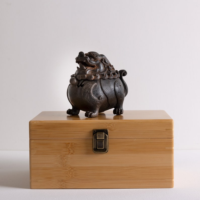 May include: A wooden box with a hinged lid and a brass latch. The box is closed and a dark brown ceramic sculpture of a mythical creature is sitting on top of the box.