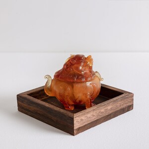 May include: A small amber-colored glass sculpture of a mythical creature, possibly a dragon or lion, with intricate details. The sculpture is sitting on a dark brown wooden tray.