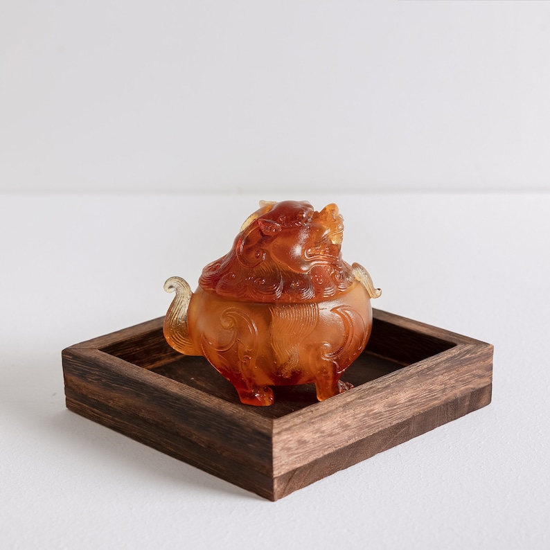 May include: A small amber-colored glass sculpture of a mythical creature, possibly a dragon or lion, with intricate details. The sculpture is sitting on a dark brown wooden tray.