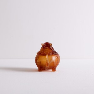 May include: A small, intricately carved amber-colored incense burner shaped like a mythical creature with a curled tail. The burner has a detailed design with intricate patterns and a lid.