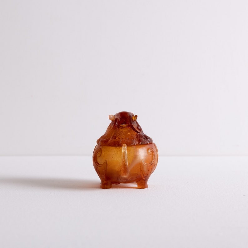 May include: A small, intricately carved amber-colored incense burner shaped like a mythical creature with a curled tail. The burner has a detailed design with intricate patterns and a lid.