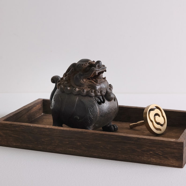 May include: A dark brown wooden tray with a black, intricately carved, lion-shaped incense burner and a gold metal incense holder shaped like a cloud.