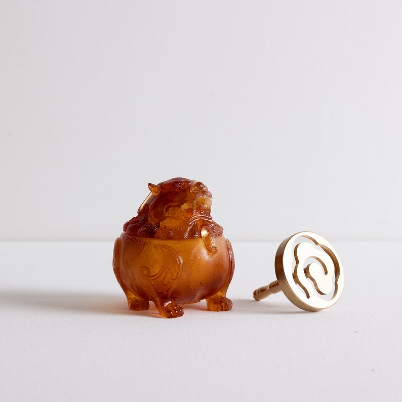 May include: A small amber-colored incense burner shaped like a mythical creature with intricate details. The burner is sitting on a white surface next to a gold-colored incense stick holder.
