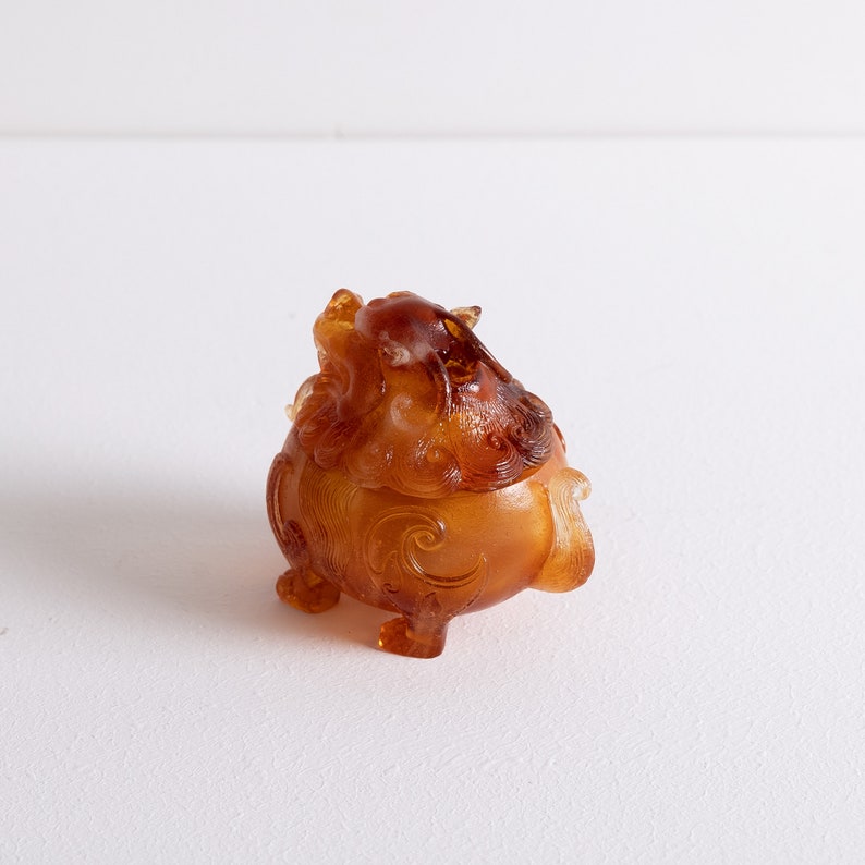 May include: A small, amber-colored, intricately carved, mythical creature figurine. The figurine is in the shape of a lion with a curled tail and detailed facial features.