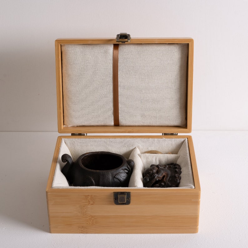 May include: A wooden box with a hinged lid, lined with fabric, and containing a black ceramic pot and a small wooden sculpture. The box is made of light brown wood and has a brass latch.