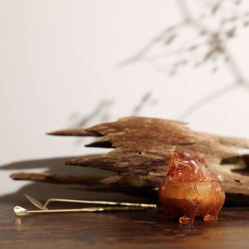 May include: A small amber-colored incense burner shaped like a mythical creature with intricate details. The burner is sitting on a piece of driftwood with a brass incense stick holder in the foreground.