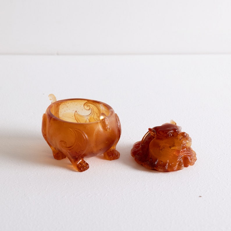 May include: A small amber-colored ceramic incense burner shaped like a dragon. The lid is separate and also shaped like a dragon. The burner has intricate details and a smooth, glossy finish.