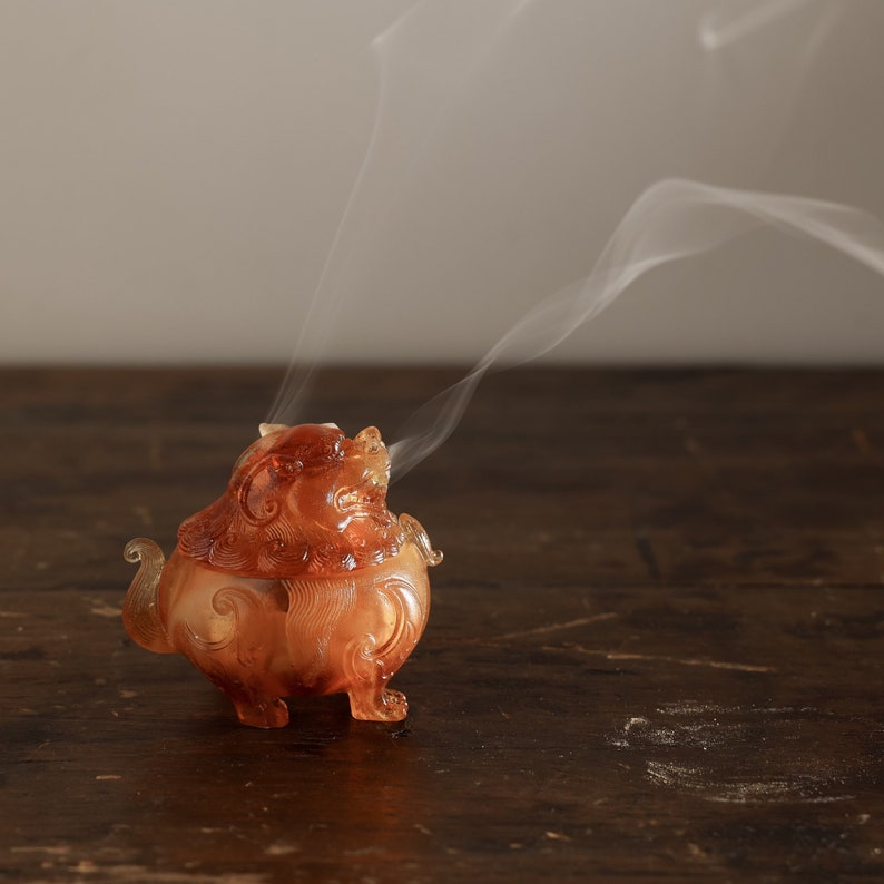 May include: A small, amber-colored incense burner shaped like a mythical creature with intricate details. Smoke curls up from the burner, which is sitting on a dark wooden surface.