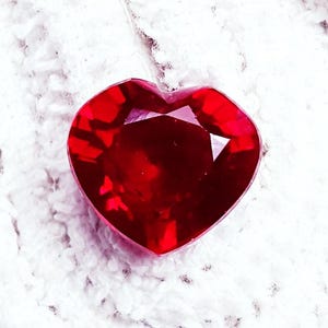 May include: A heart-shaped, faceted, deep red gemstone. The gem is set against a white, textured background. The cut of the stone creates multiple facets that reflect light, giving it a brilliant appearance. The gemstone's shape is a classic symbol of love.