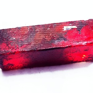 May include: A rectangular, deep red gemstone with a rough, textured surface. The stone has a dark, almost black, base colour with streaks of red and brown. The edges are uneven, giving it a natural, raw appearance.