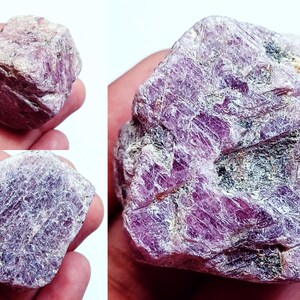 May include: A raw, unpolished ruby gemstone with a deep magenta and purple hue. The stone features a rough, crystalline texture with visible natural facets, internal fractures, and dark mineral inclusions throughout its irregular, blocky shape.