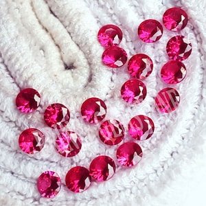 May include: A collection of small, round, faceted loose gemstones in a vibrant, deep pinkish-red colour. Each stone features a brilliant cut with multiple reflective surfaces that catch the light, creating a sparkling appearance across the group.