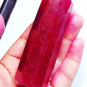 May include: A faceted, rectangular ruby gemstone with a deep red colour. The gemstone is held in a hand, showcasing its polished surfaces and natural imperfections. The ruby's shape is a long, symmetrical prism.