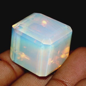 Puede incluir: Una piedra preciosa de ópalo rectangular y facetada, de aspecto lechoso e iridiscente. La gema muestra un juego de colores con destellos de azul, amarillo y naranja. La piedra se sostiene en una mano sobre un fondo oscuro.