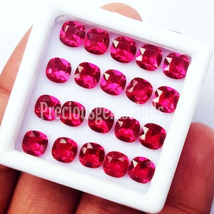 May include: A collection of nineteen cushion-cut, vibrant pinkish-red gemstones arranged in a grid within a white square display box. The stones feature faceted surfaces that reflect light, and the text Preciousgemshinds is visible in white.