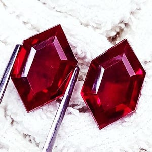 May include: Two loose, faceted gemstones with a deep red color and a distinct kite-like geometric shape. The stones feature sharp, angular edges and a polished surface that reflects light, showcasing a vibrant, saturated crimson hue throughout.