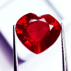 May include: A heart-shaped, faceted, deep red gemstone is held by a pair of silver tweezers against a white background. The gemstone's facets catch the light, creating a sparkling effect. This gemstone is likely a ruby, perfect for jewelry.