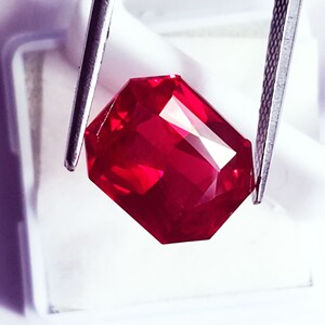 May include: A vibrant, deep red gemstone with a rectangular emerald cut, held by a pair of silver-toned metal tweezers. The stone features multiple facets that reflect light, creating a brilliant, clear appearance with intense crimson color saturation.