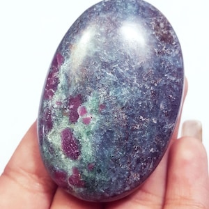 Op de afbeelding: Een ovaalvormige gepolijste edelsteen met een gevlekt oppervlak met diep magenta-rode insluitingen tegen een donkerblauw-grijze en lichtgroene matrix. De steen heeft een gladde, glanzende afwerking met overal zichtbare kristallijne texturen.