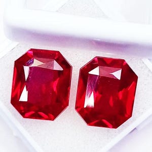 May include: Two vibrant, deep red gemstones with an emerald-cut shape, featuring multiple facets that reflect light. These two loose stones are displayed side-by-side within a white, rectangular storage box with a foam-lined interior base.